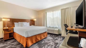 Habitación de hotel con cama, mesita de noche, lámpara, televisor, escritorio, silla y ventana con cortinas.