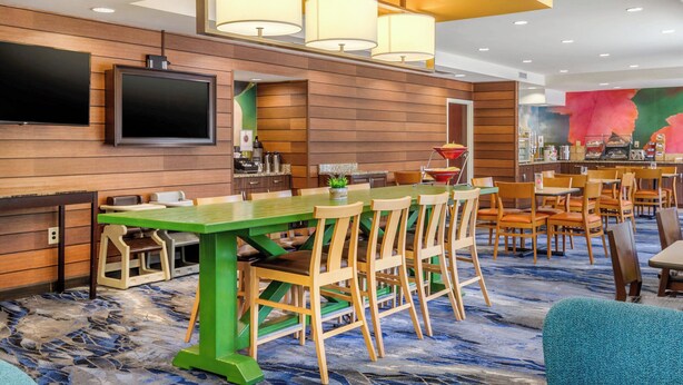 A hotel lobby with a long green table, wooden chairs, a flat-screen TV, and a colorful mural. - Fairfield Inn by Marriott Myrtle Beach North (Myrtle Beach)