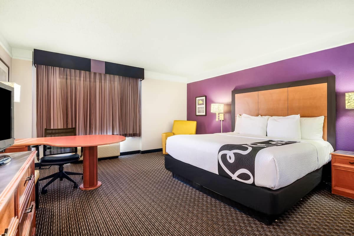 A hotel room with a bed, a desk, a chair, a nightstand, and a window with curtains.