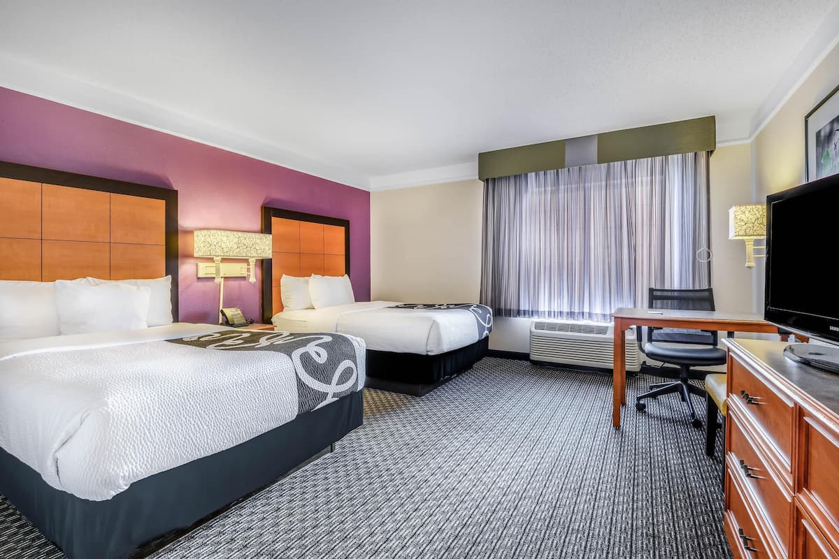 A hotel room with two beds, a desk, a TV, and a window with curtains.