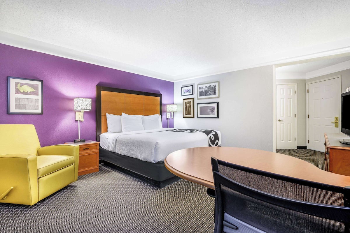 A hotel room with a bed, a yellow armchair, a small table, and a desk.
