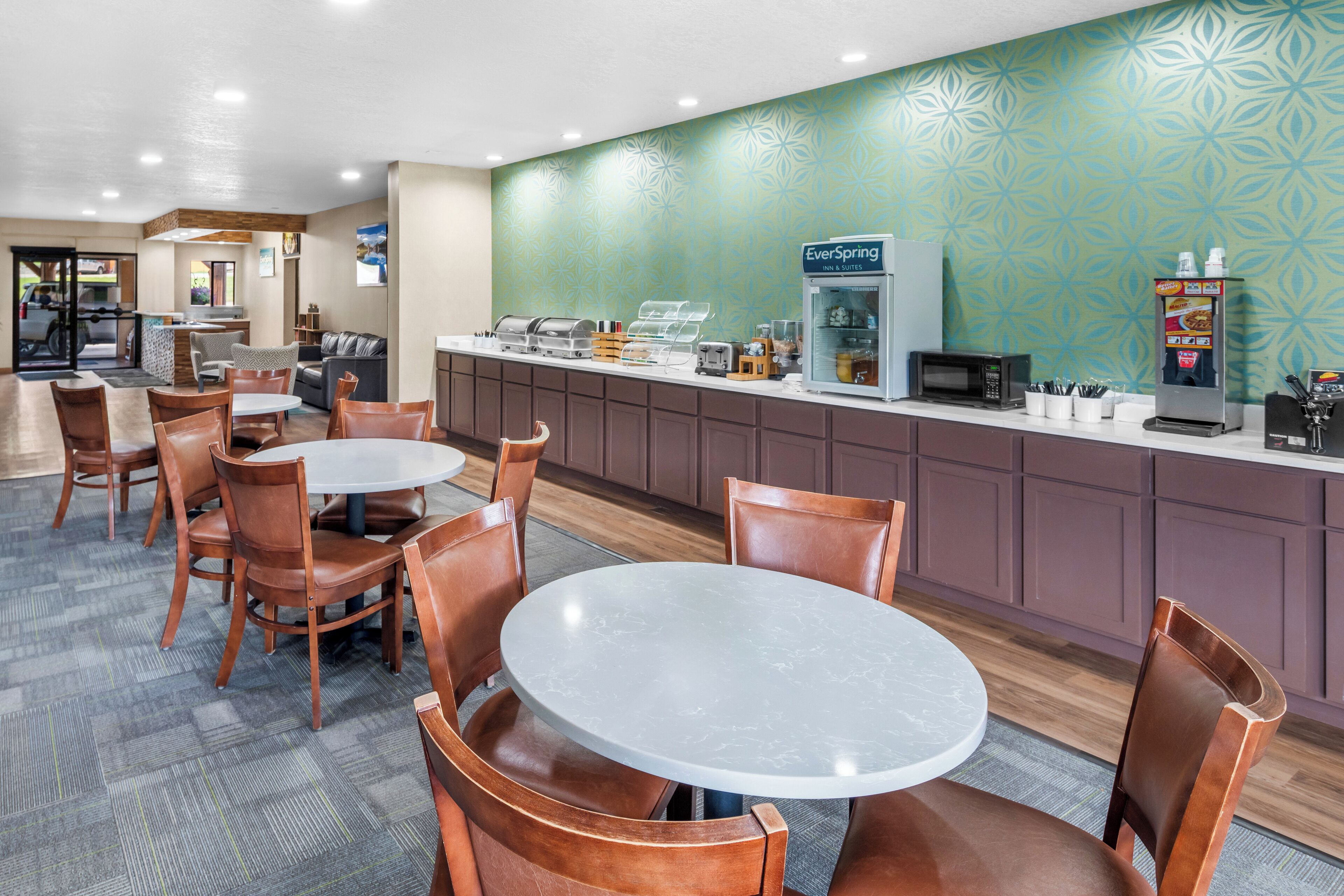 Eine Hotellobby mit einer Sitzecke, die runde Tische und Stühle, eine Kaffeestation mit einer beschrifteten Kaffeemaschine und einer gemusterten Wand umfasst.