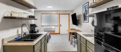 A compact kitchen with a sink, countertop, wooden cabinets, a dining table, and a TV mounted on the wall.