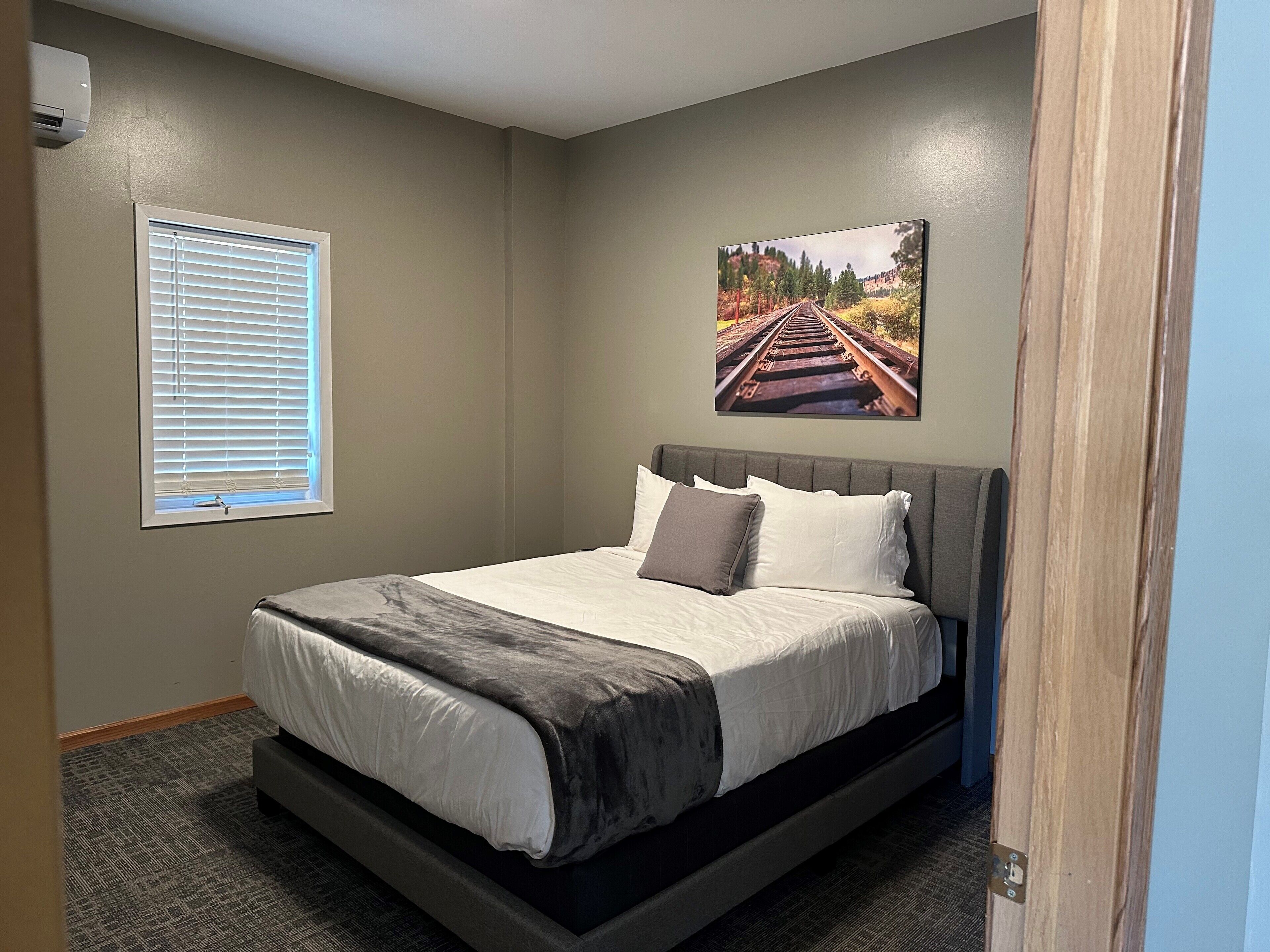 A bedroom with a bed, a window with blinds, a wall-mounted air conditioner, and a picture of a railway track.