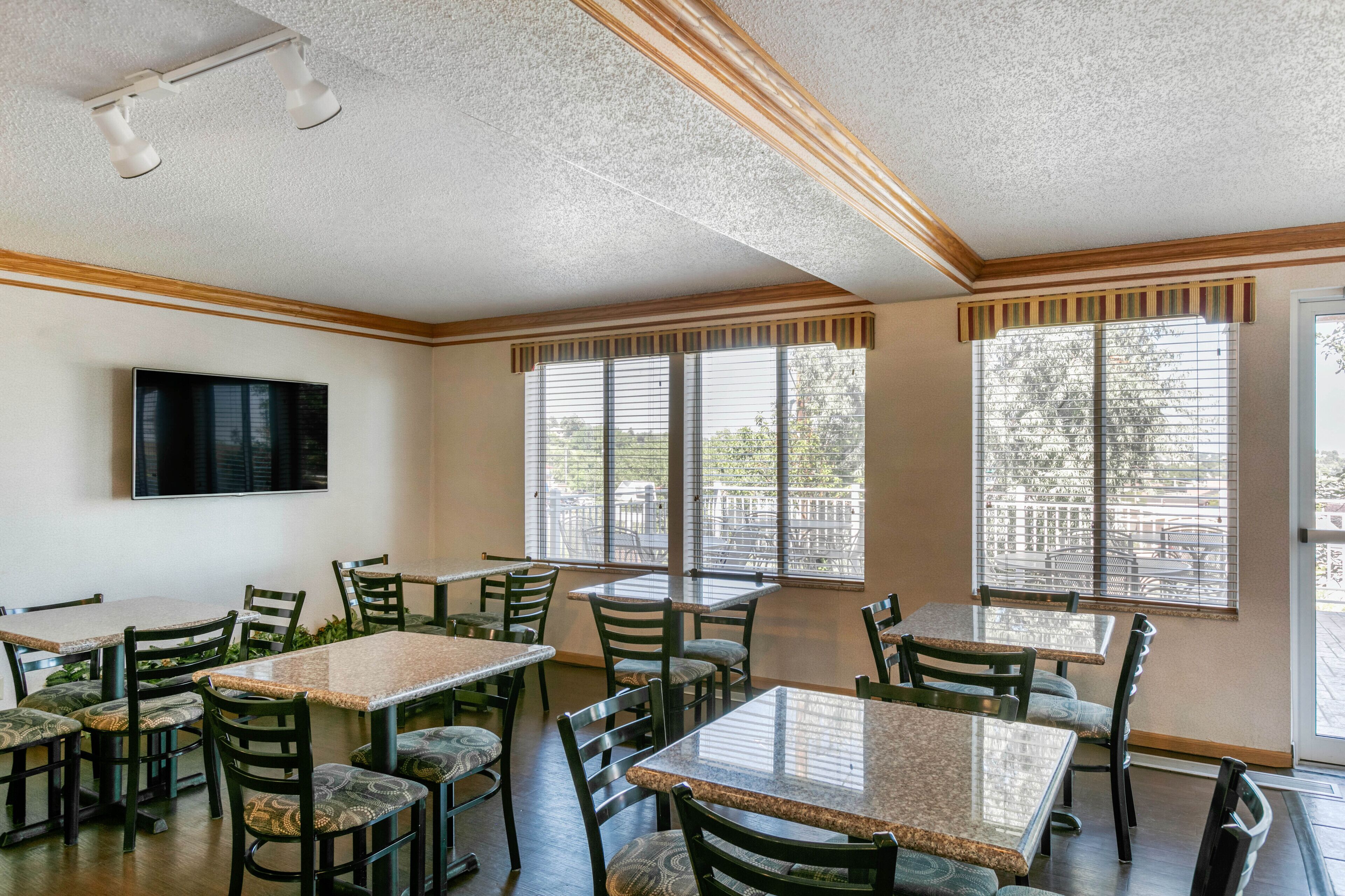 A dining area with multiple tables and chairs, large windows with blinds, and a flat-screen TV mounted on the wall.