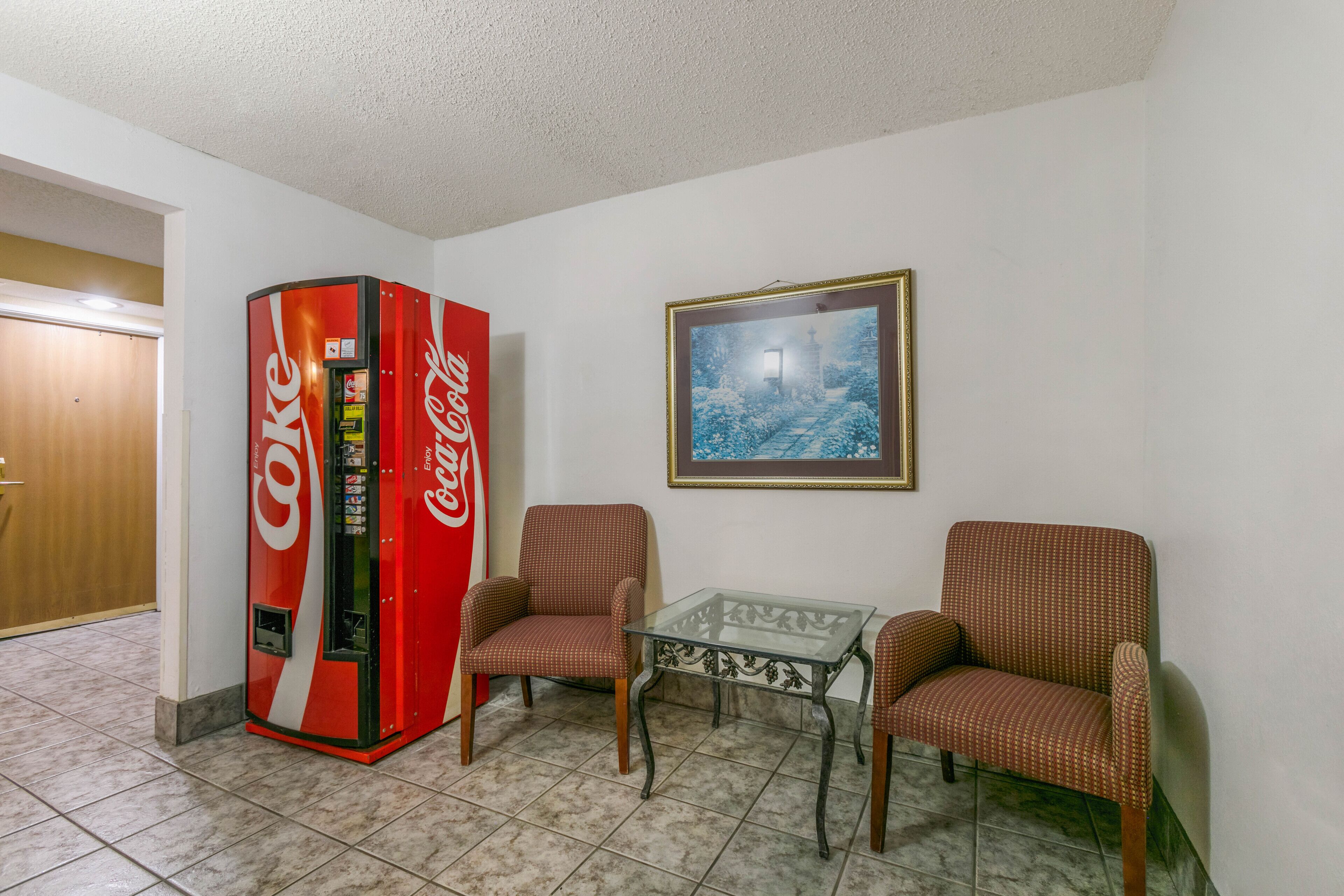 Una habitación con una máquina expendedora de Coca-Cola, dos sillas, una mesa de vidrio y un cuadro en la pared.