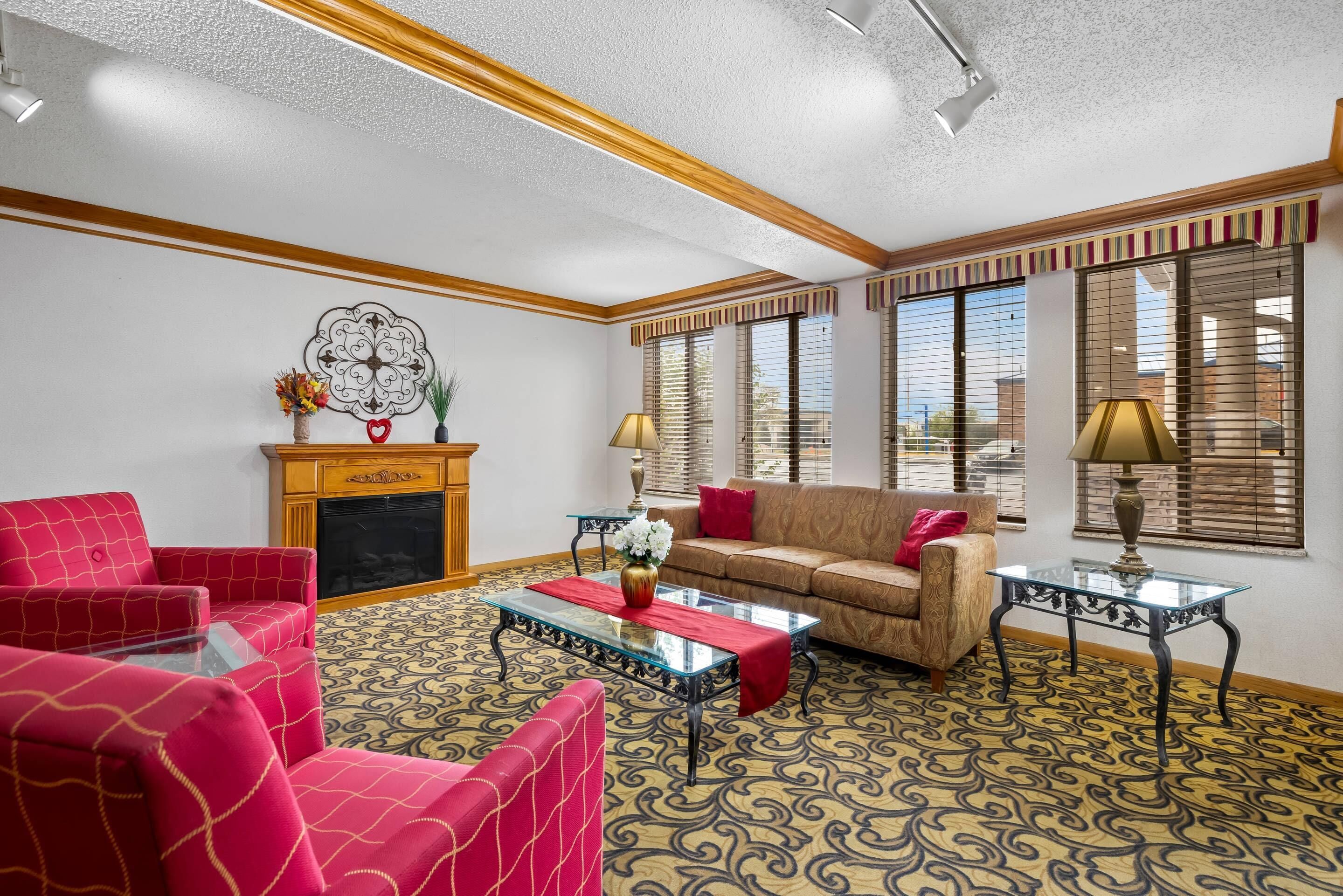 A living room with a fireplace, red and beige sofas, patterned carpet, and large windows with blinds.