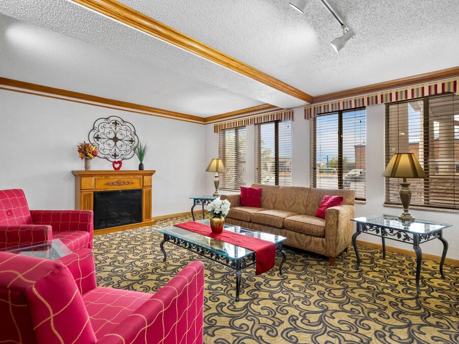 A living room with a fireplace, red and beige sofas, patterned carpet, and large windows with blinds.