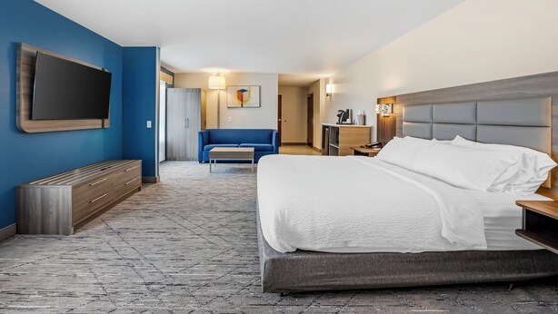 A modern hotel room with a large bed, a flat-screen TV mounted on the wall, a blue sofa, and a small desk with a lamp.