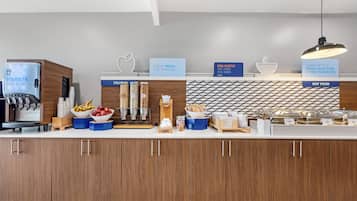 A modern breakfast bar with a coffee machine, fruit bowls, and labeled containers for hot food.
