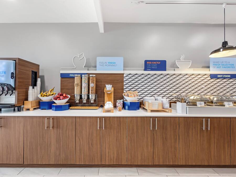 A modern breakfast bar with a coffee machine, fruit bowls, and labeled containers for hot food.