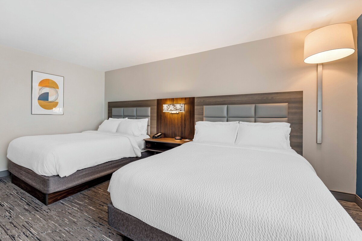 A hotel room with two beds, a nightstand with a lamp, and a wall-mounted phone.