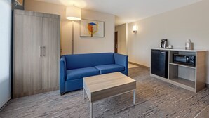 A modern room with a blue sofa, a wooden coffee table, a wardrobe, a kitchenette with a fridge and microwave, and a painting on the wall.