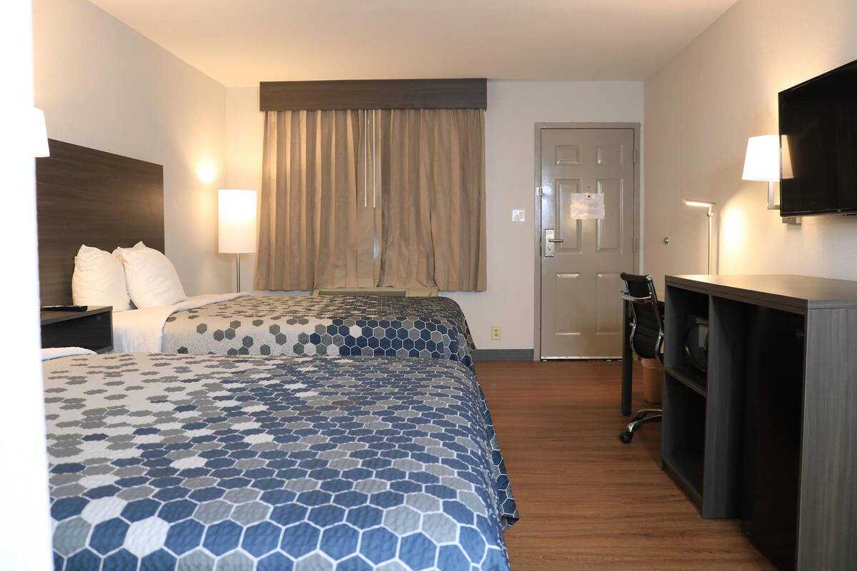 A hotel room with two beds, a desk, a chair, a TV, and a door.