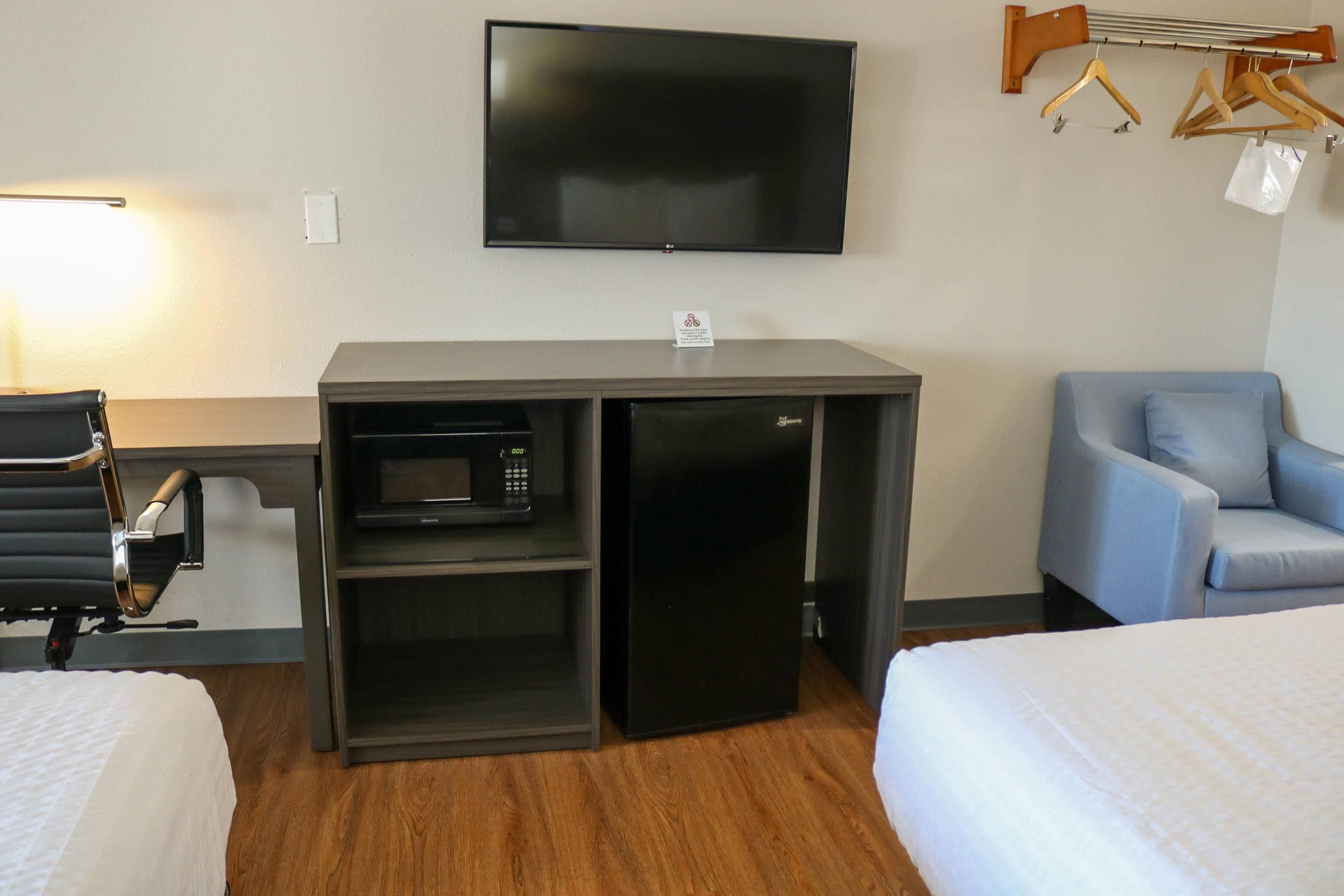 A hotel room with a flat-screen TV mounted on the wall, a microwave in a cabinet, a desk with a chair, and a sofa.