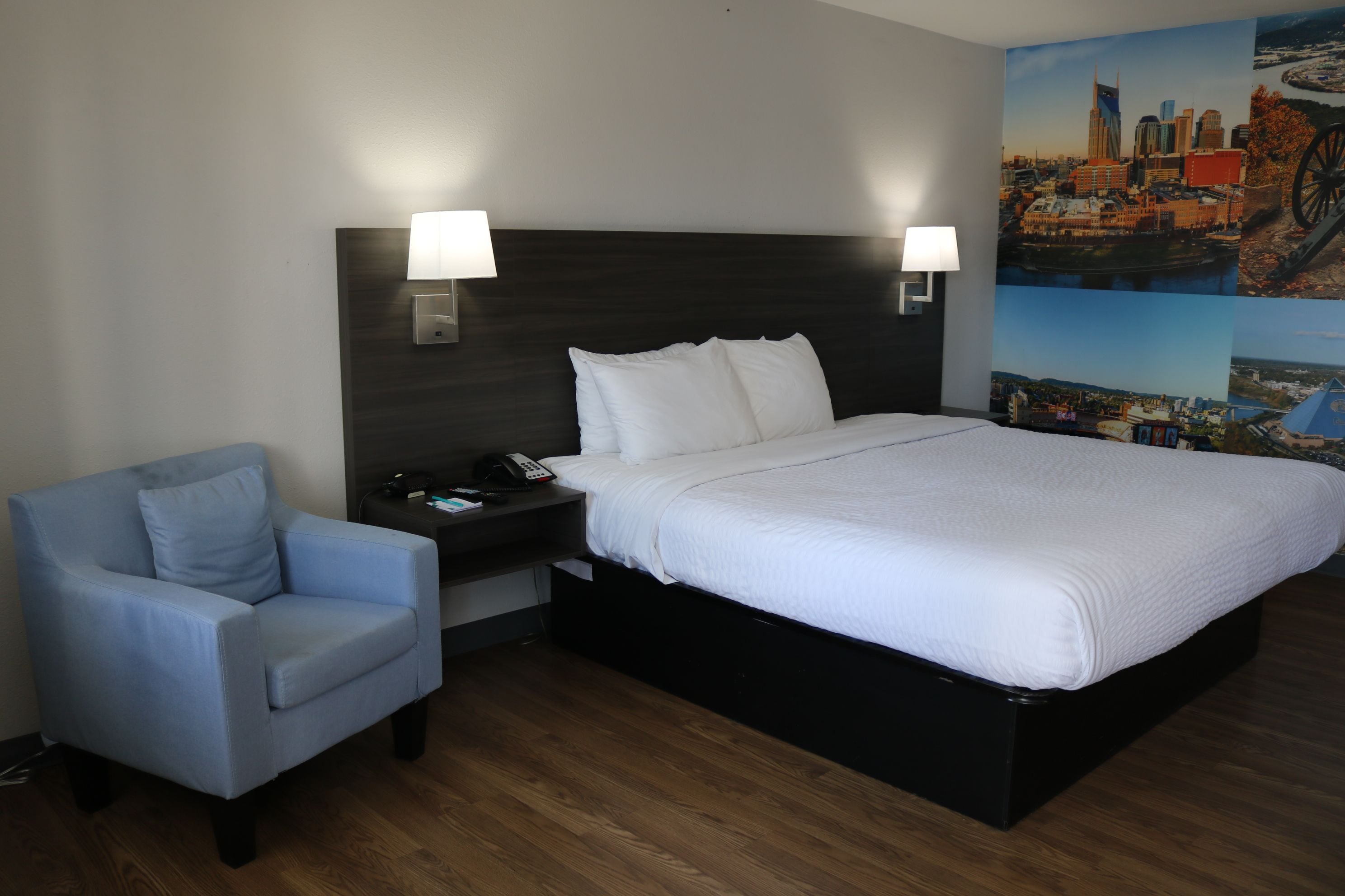 A hotel room with a bed, a chair, a nightstand, and a wall with a cityscape mural.
