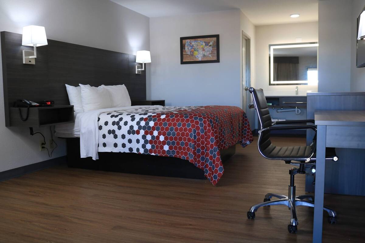 A hotel room with a bed, desk, chair, and a bathroom mirror.
