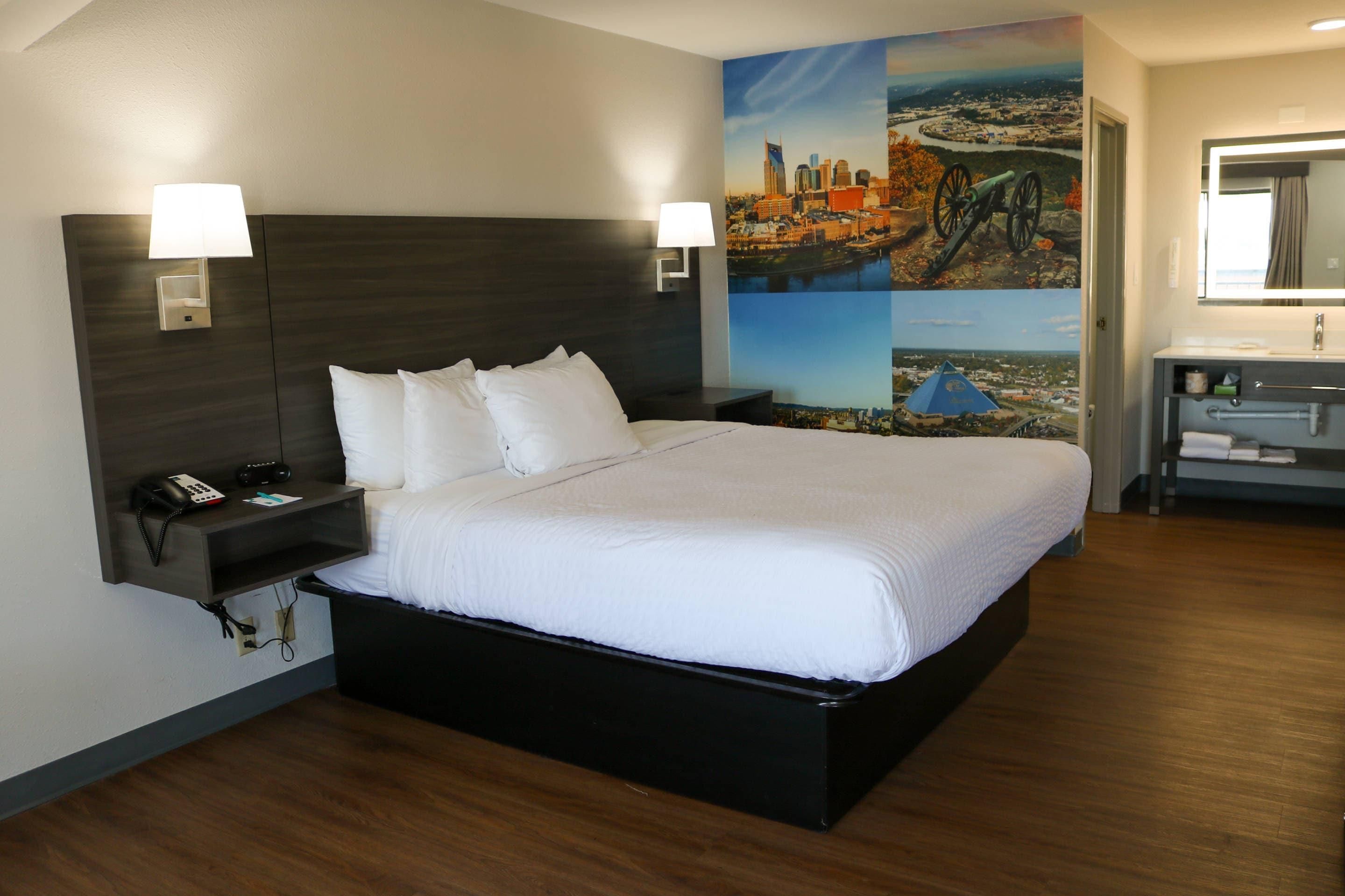 Habitación de hotel con una cama grande, lámparas de noche, una mesita de noche con teléfono y radio, baño con lavabo y espejo, y un mural con un paisaje urbano en la pared.
