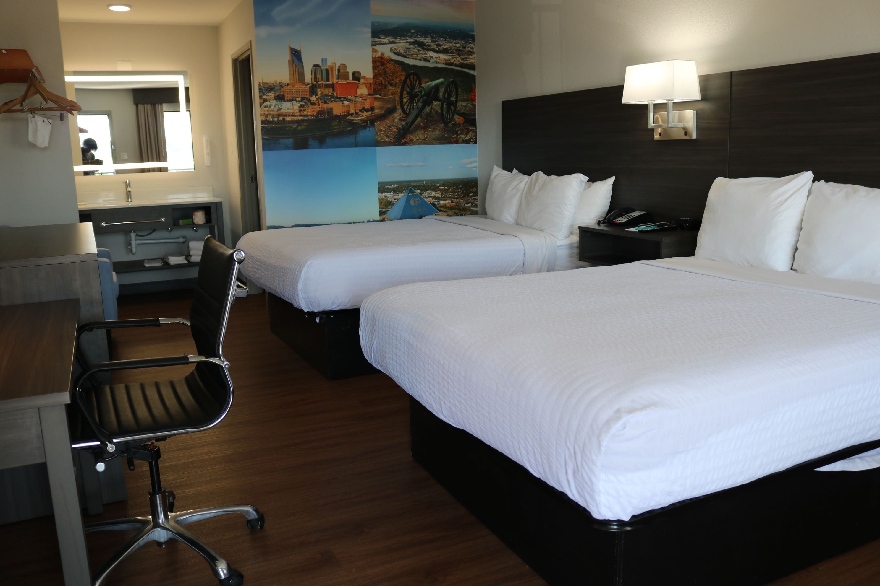 A hotel room with two beds, a desk, a chair, a lamp, and a painting on the wall.