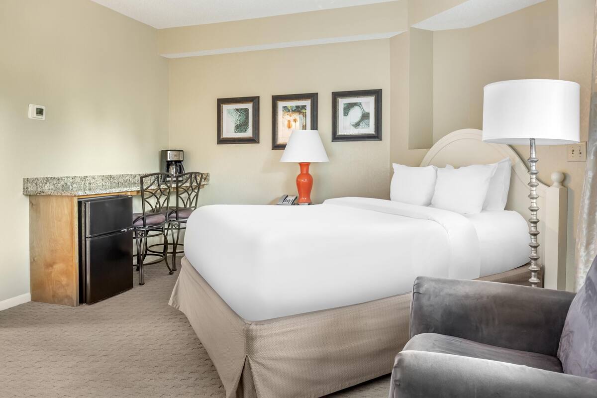 A hotel room with a bed, a desk with a coffee maker, a chair, and a lamp.