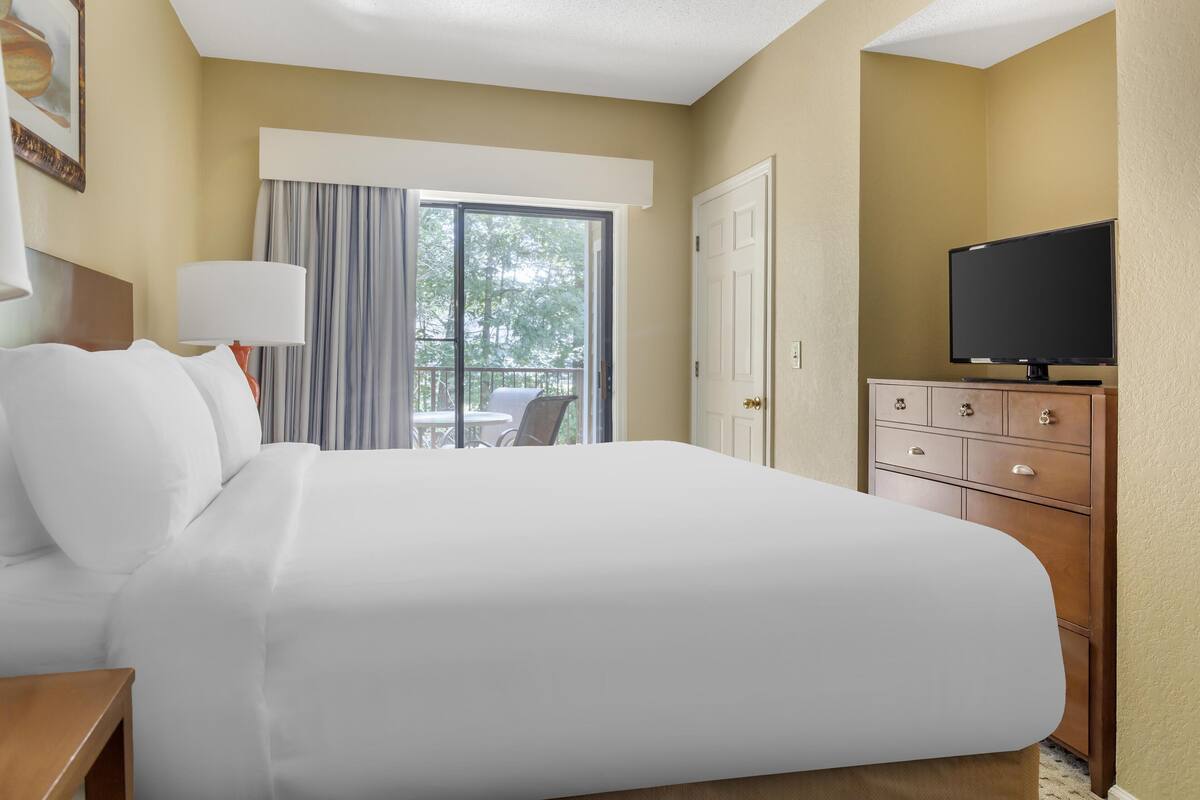 A hotel room with a large bed, a television on a wooden dresser, and a balcony with a table and chairs.