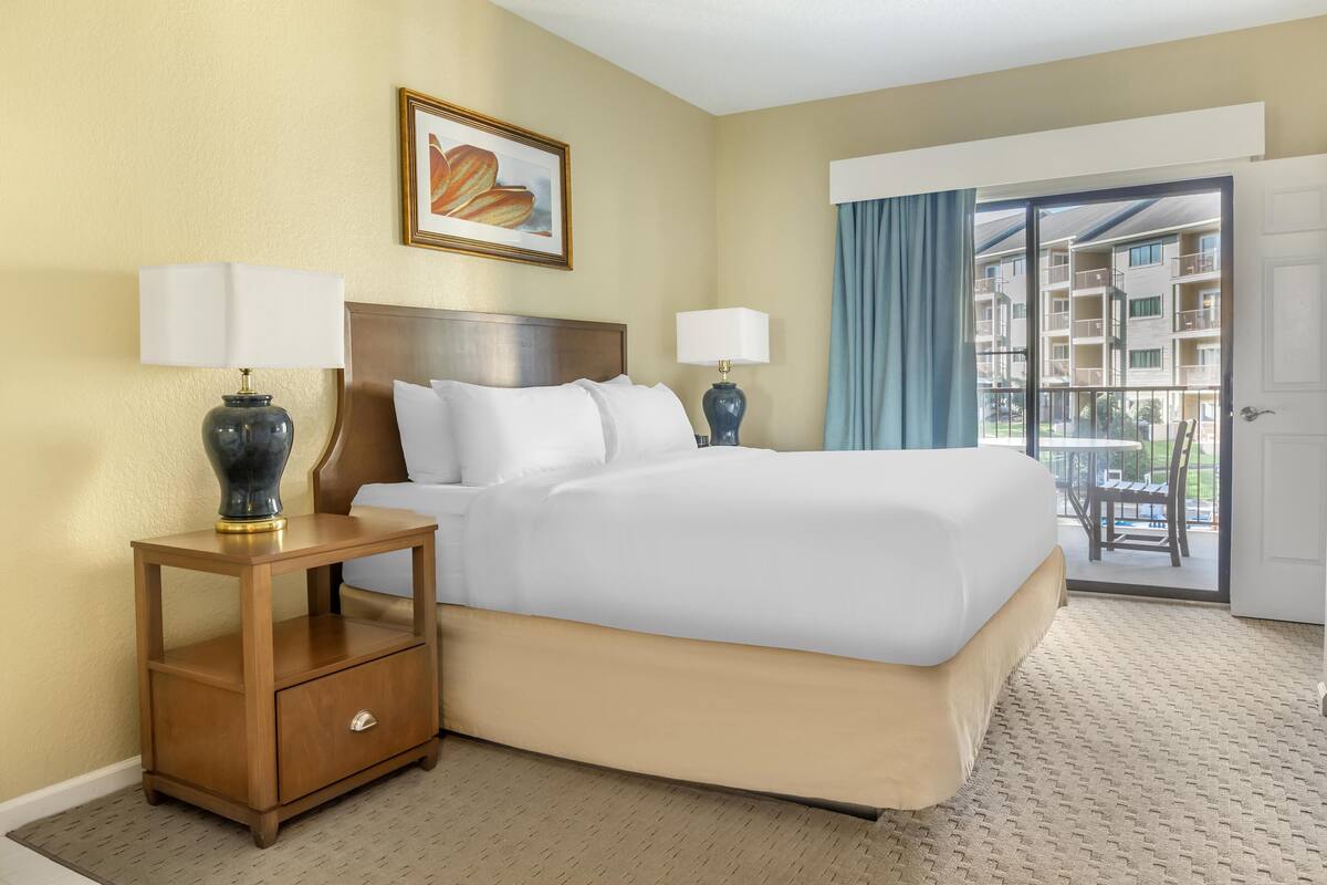 A hotel room with a large bed, two bedside tables with lamps, a framed picture on the wall, and a view of the outside balcony.
