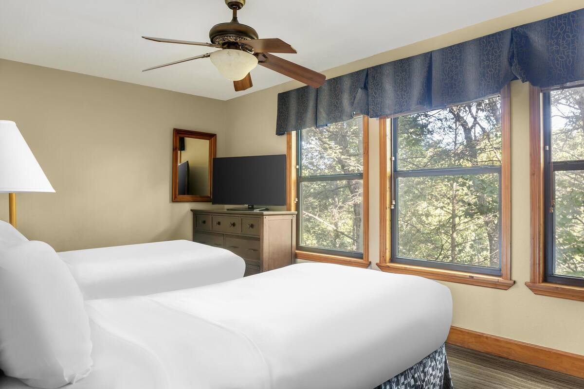 A hotel room with two beds, a ceiling fan, a television on a wooden dresser, and large windows with a view of trees.