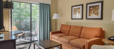 A living room with a sofa, a coffee table, and a striped armchair. There is a window with curtains, a lamp, and two framed pictures on the wall.