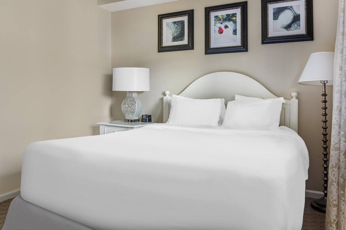 A neatly made bed with white linens, a bedside table with a lamp, and framed artwork on the wall.