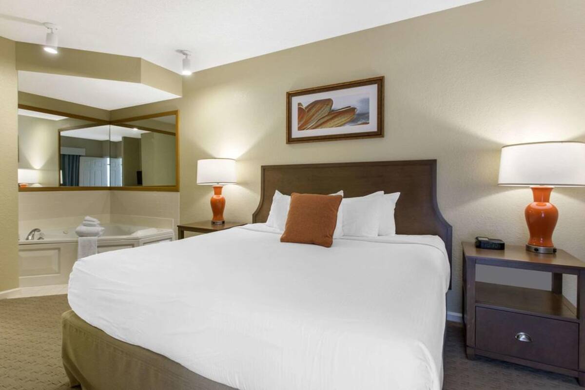A hotel room with a large bed, two bedside tables with lamps, a mirror, a painting on the wall, and a bathroom with a bathtub and sink.