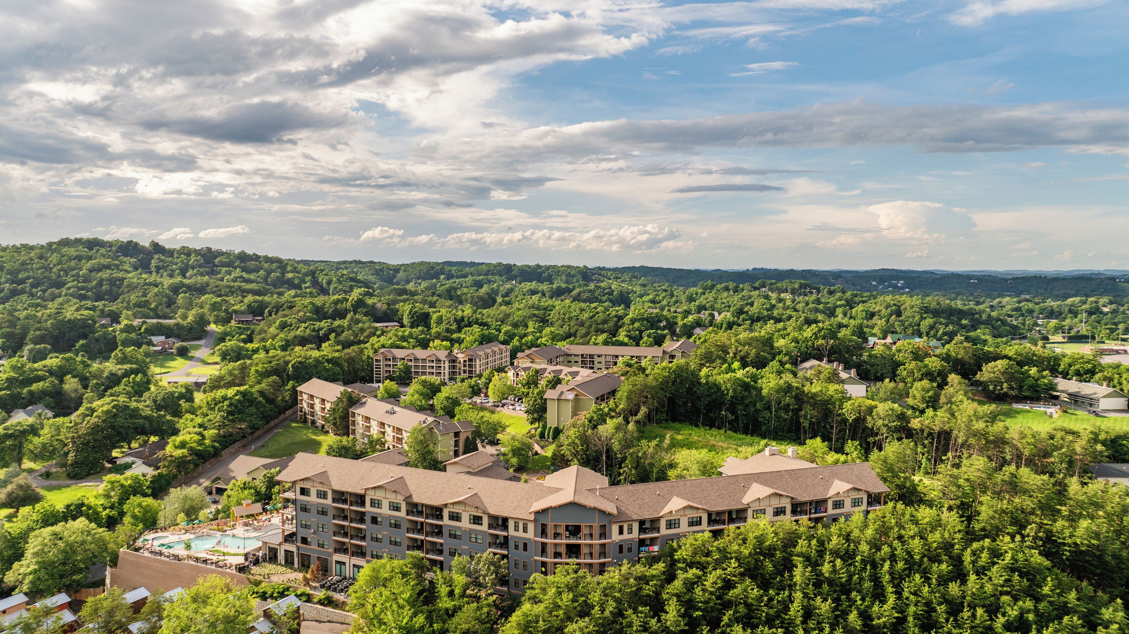 A resort with multiple buildings surrounded by dense forest, featuring a swimming pool and guest rooms.