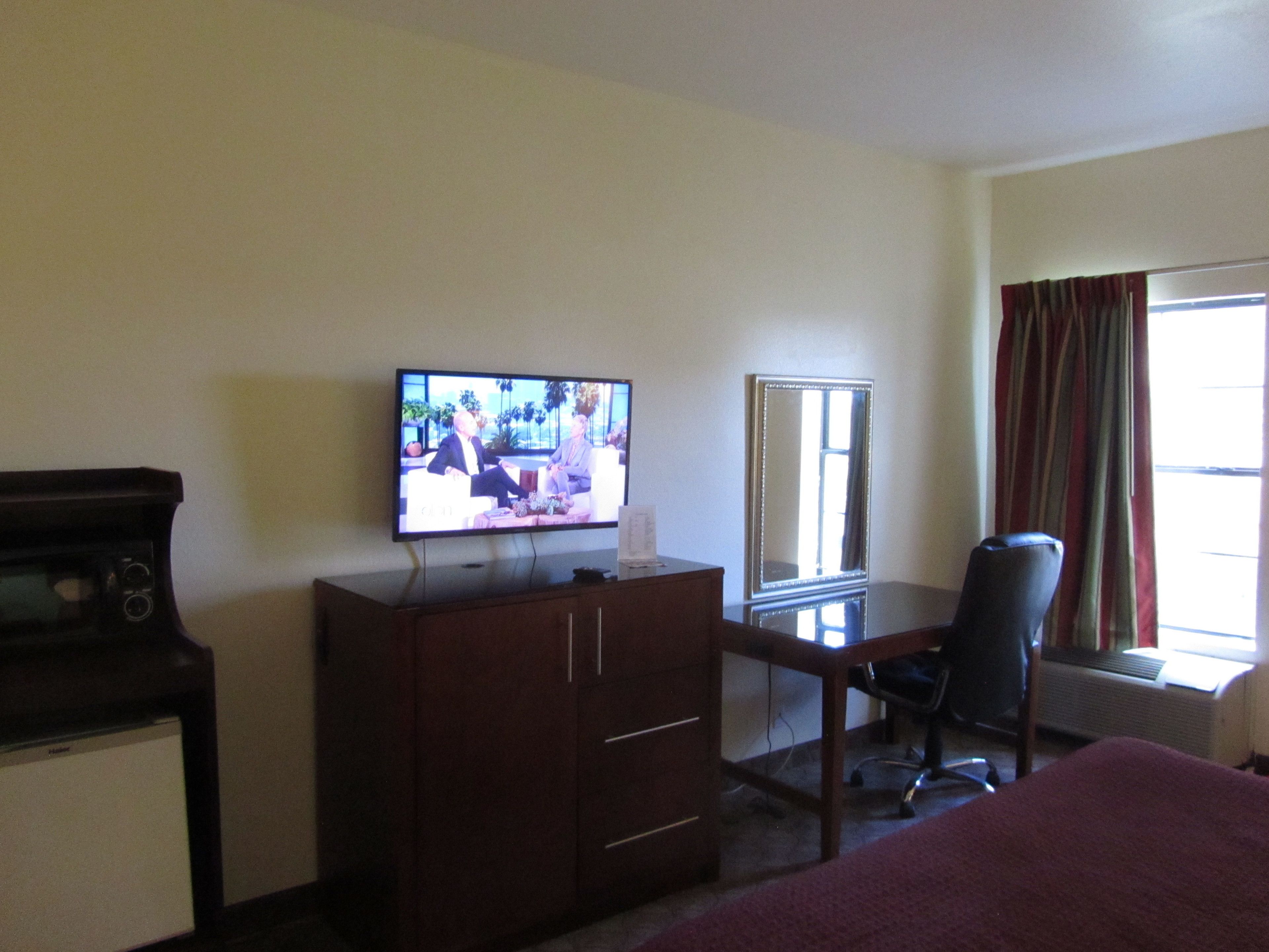 A hotel room with a television on a wooden dresser, a desk with a chair, a large mirror, and a window with curtains.