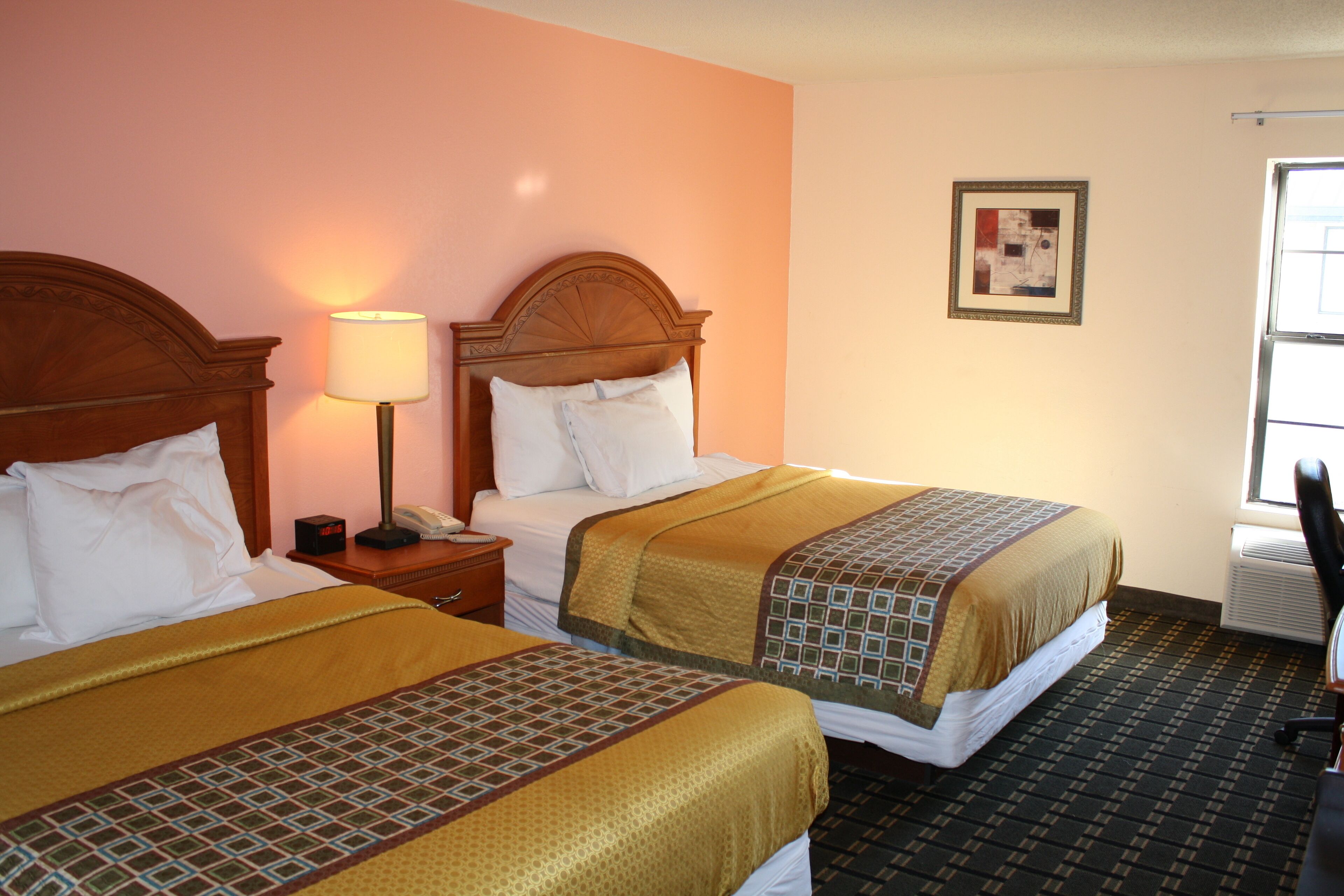 A hotel room with two beds, a nightstand with a lamp, a framed picture on the wall, and an air conditioning unit.