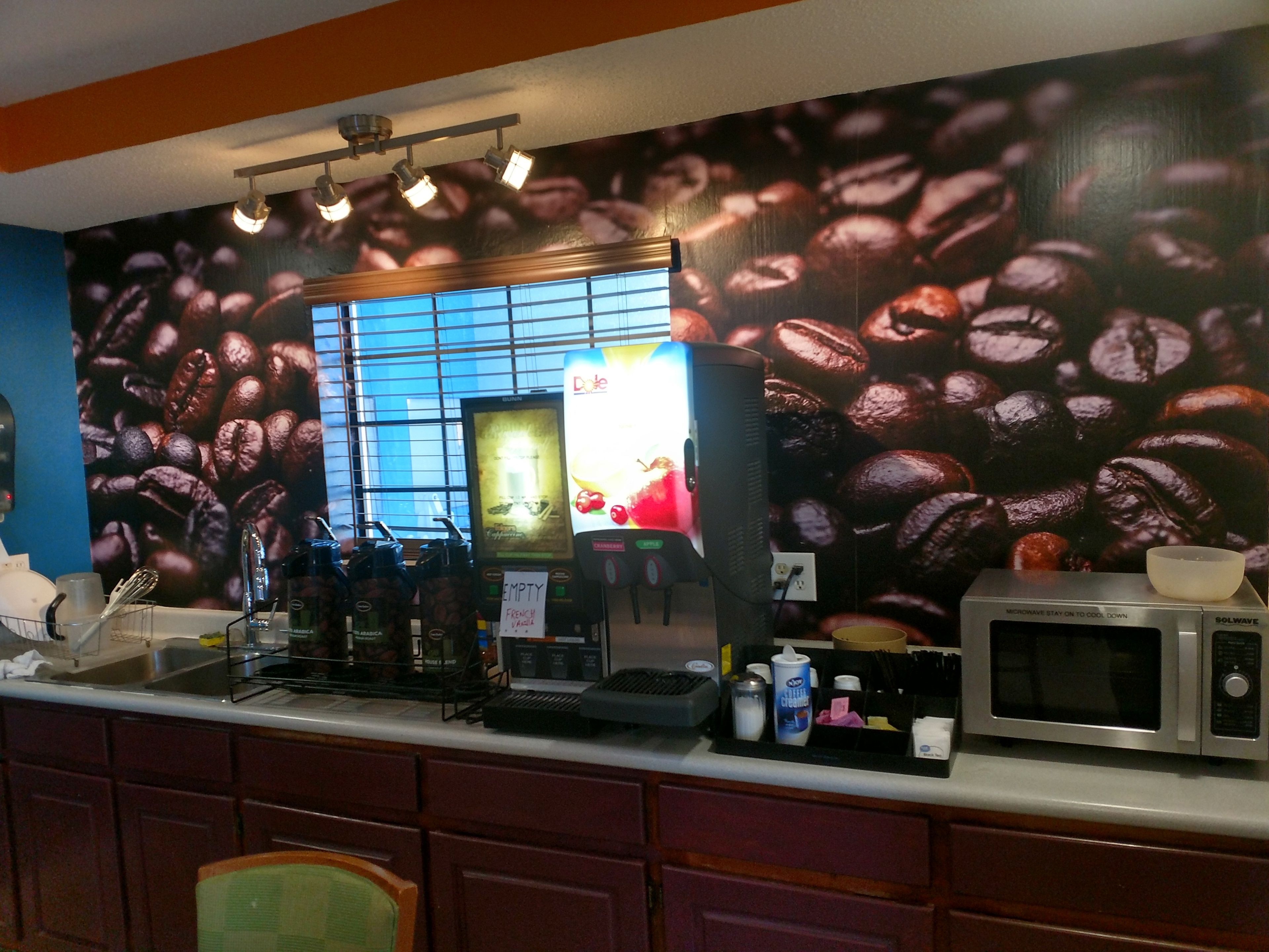 Una cafetería con un mural de granos de café, una máquina de café, un microondas y una pizarra de menú.