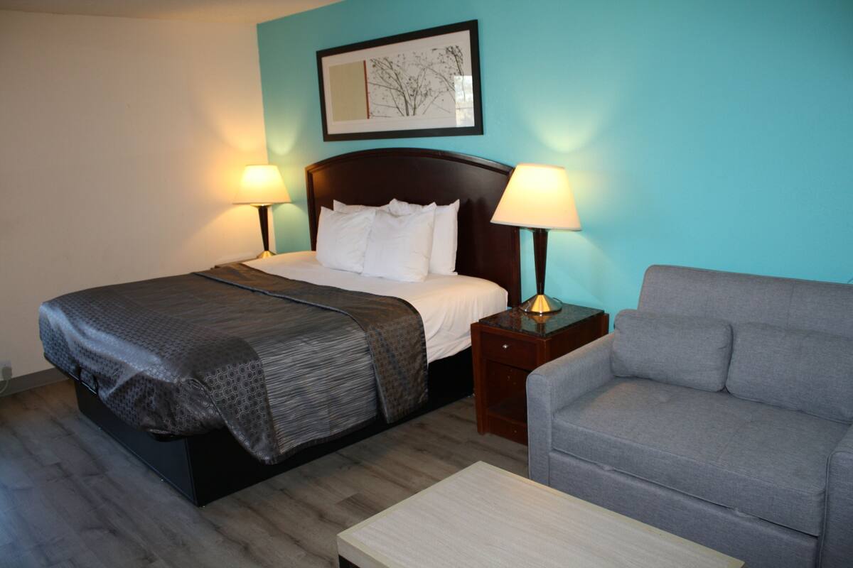 A hotel room with a bed, two bedside lamps, a gray sofa, and a wooden coffee table.