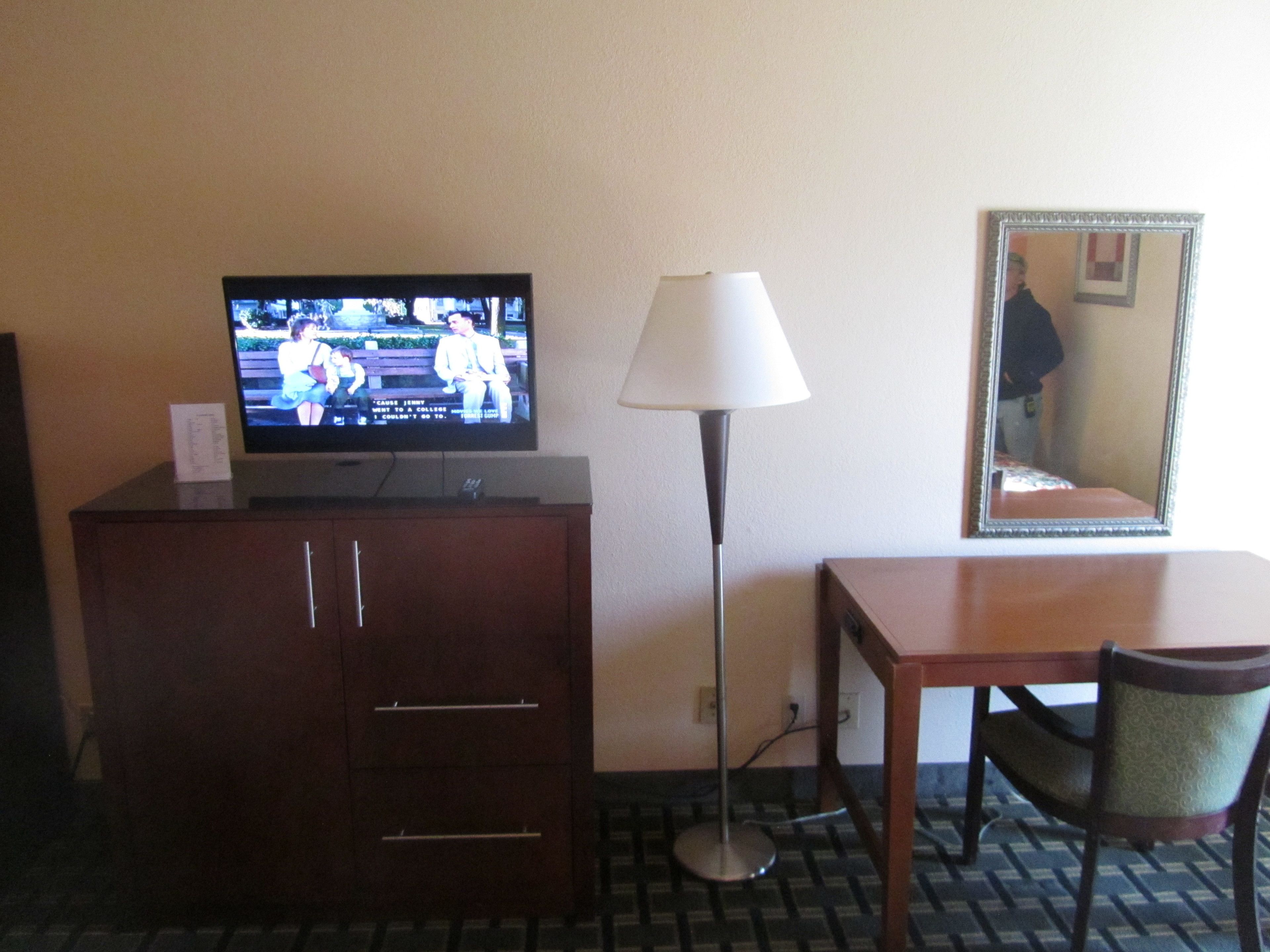 A hotel room with a TV on a wooden cabinet, a lamp, a mirror, and a table with a chair.