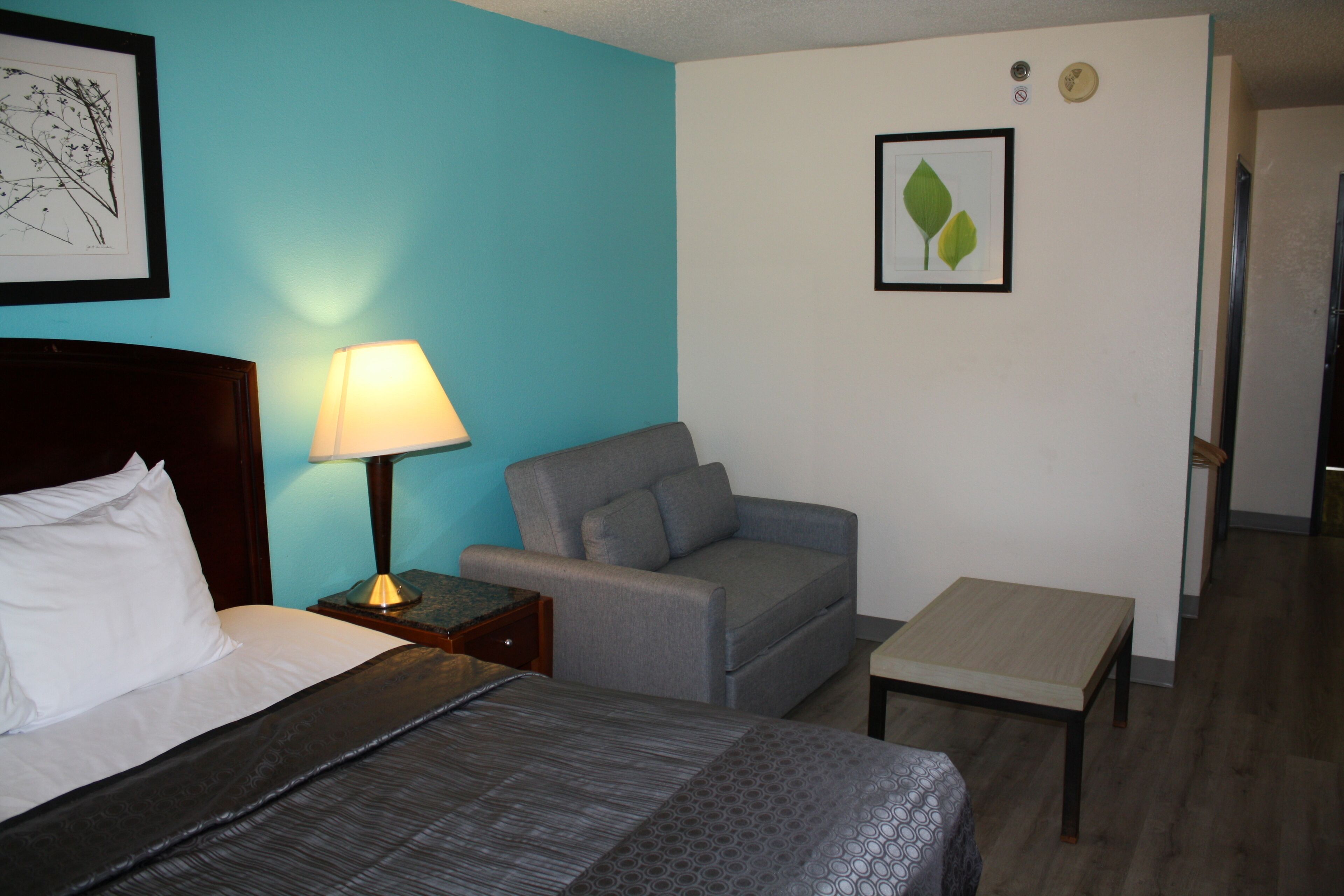 A hotel room with a bed, a grey sofa, a small table, a lamp, and two framed pictures on the walls.