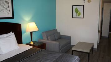A hotel room with a bed, a grey sofa, a small table, a lamp, and two framed pictures on the walls.
