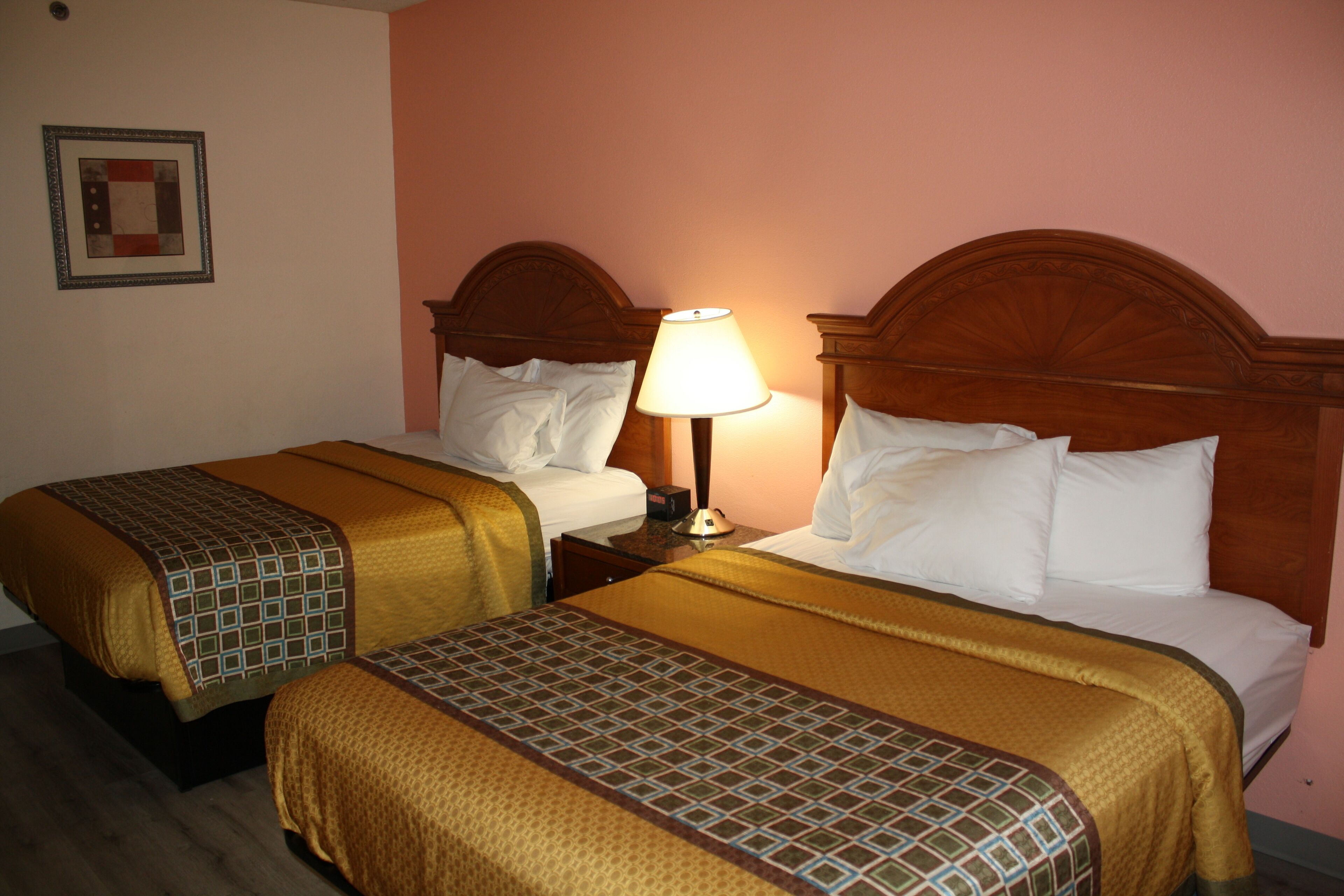 Habitación de hotel con dos camas, cada una con cabecero de madera, mesita de noche con lámpara y un cuadro en la pared.