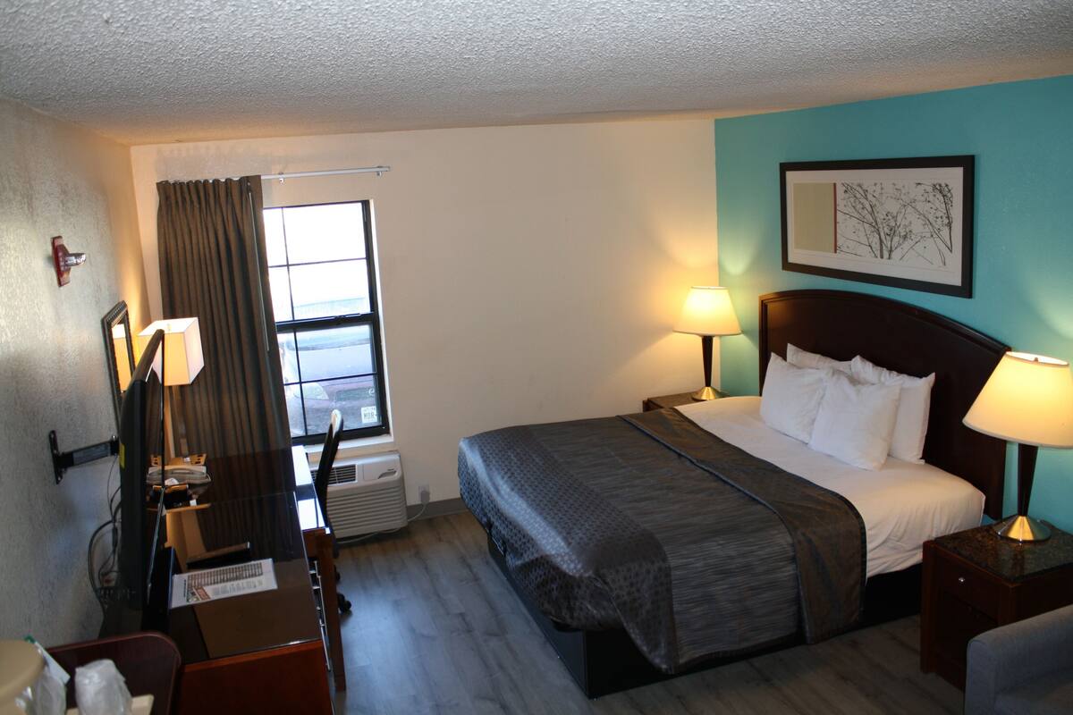 A hotel room with a bed, two bedside lamps, a window with curtains, a desk with a chair, and a wall-mounted picture.