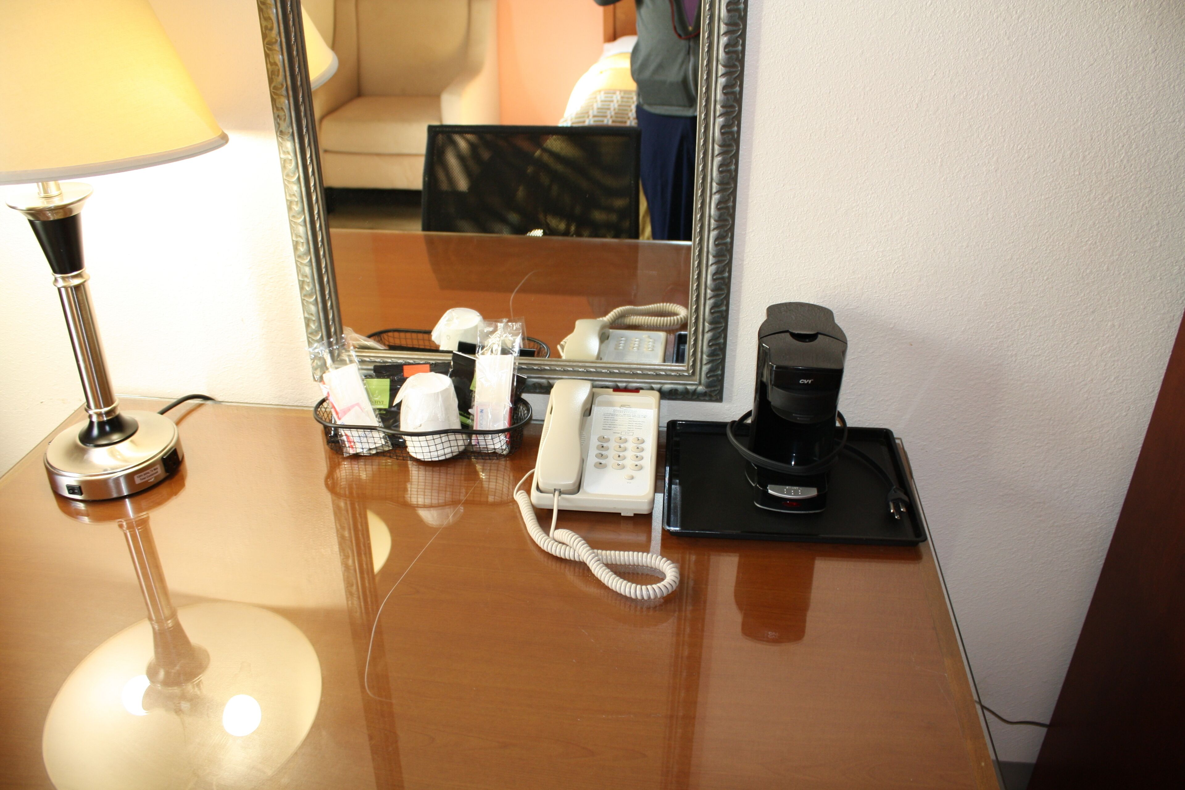 Una camera d'albergo con una scrivania in legno, una lampada, un telefono, una macchina del caffè e uno specchio che riflette l'ambiente.