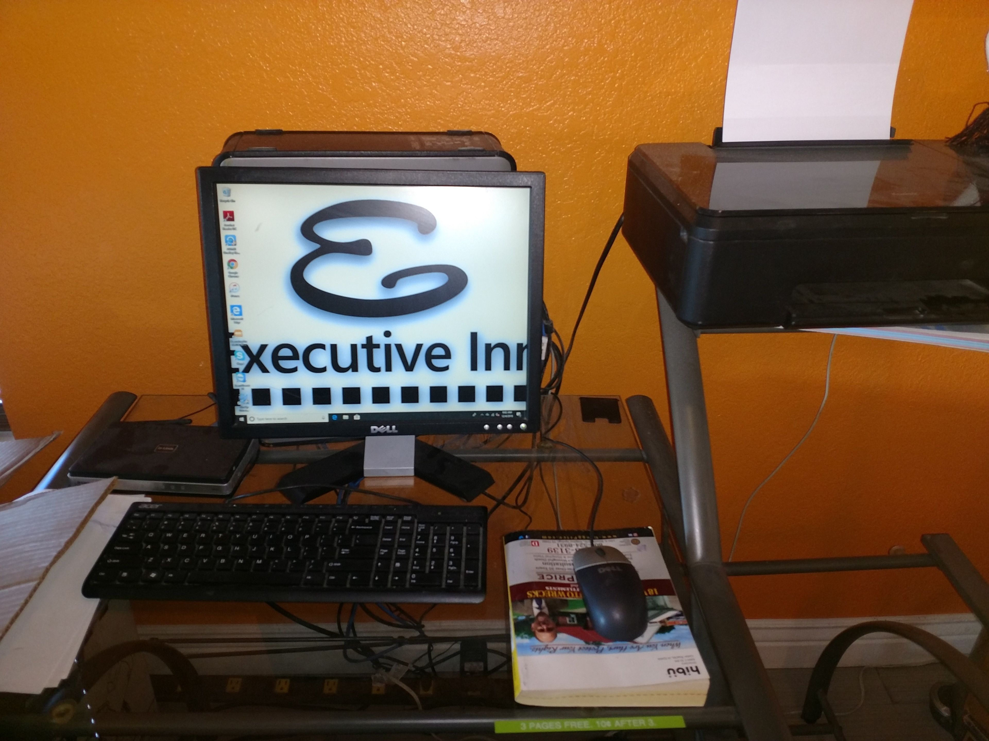 A computer setup with a monitor displaying 'Executive Intranet', a keyboard, a mouse, and a book on a desk against an orange wall.