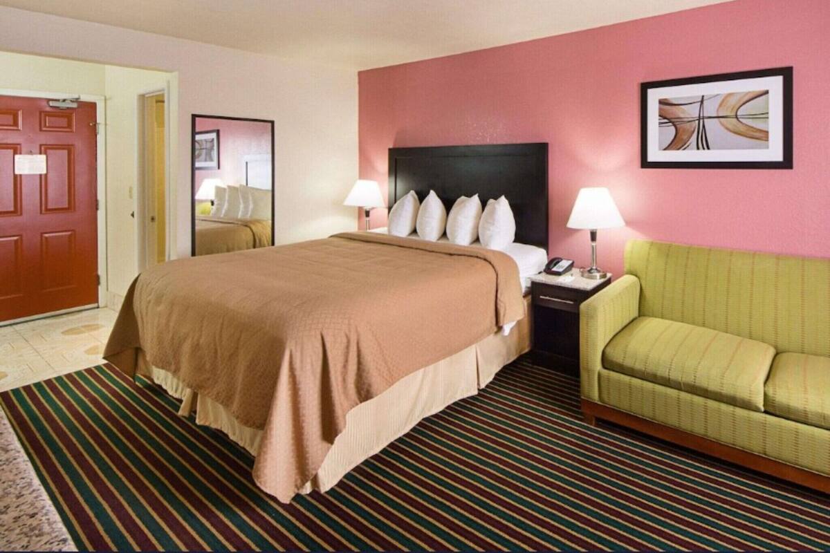 A hotel room with a bed, bedside tables, a green armchair, a door, and a framed picture on the wall.