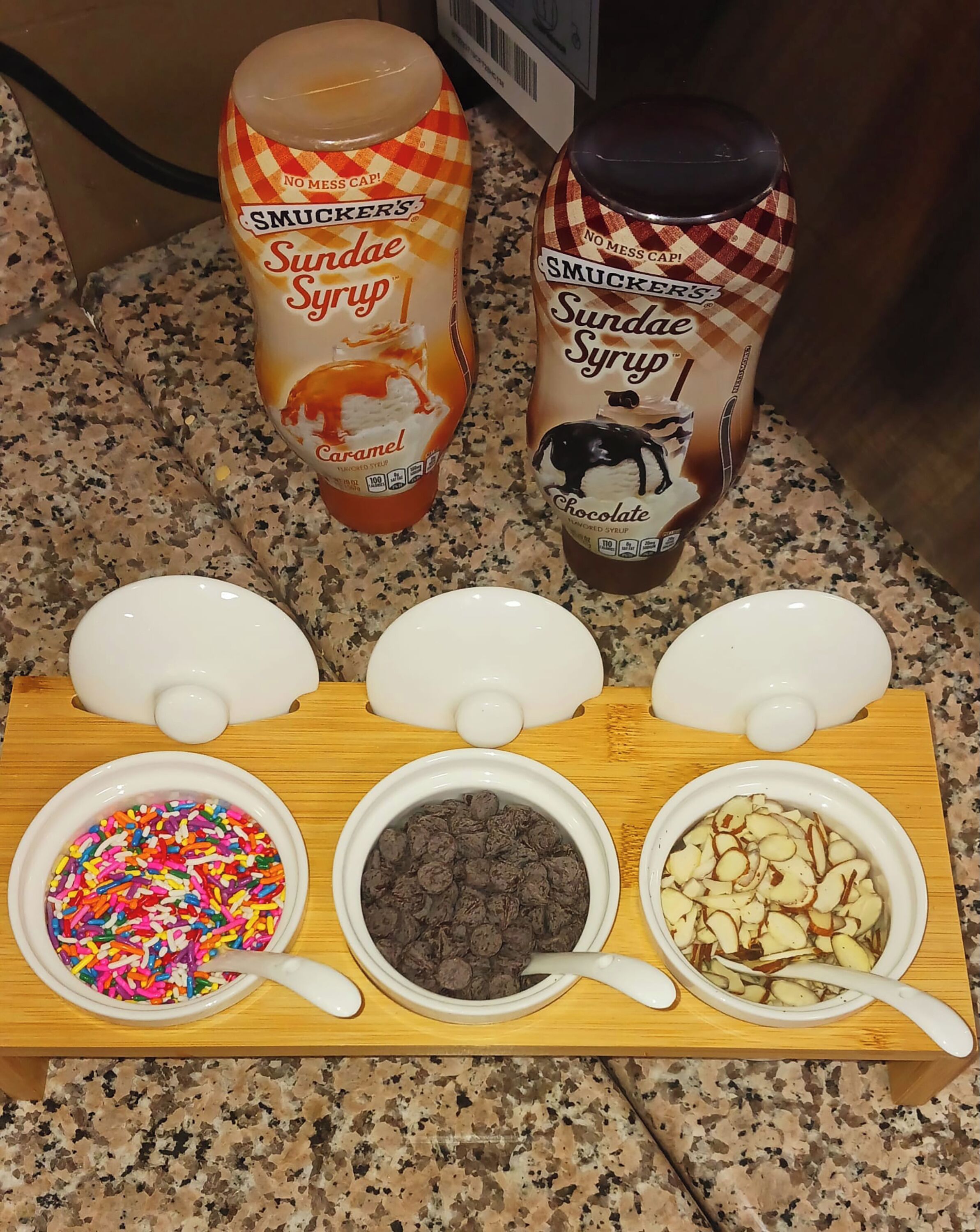 A kitchen counter with two bottles of Smucker's Sundae Syrup, one caramel and one chocolate, and three bowls containing sprinkles, chocolate chips, and sliced almonds.