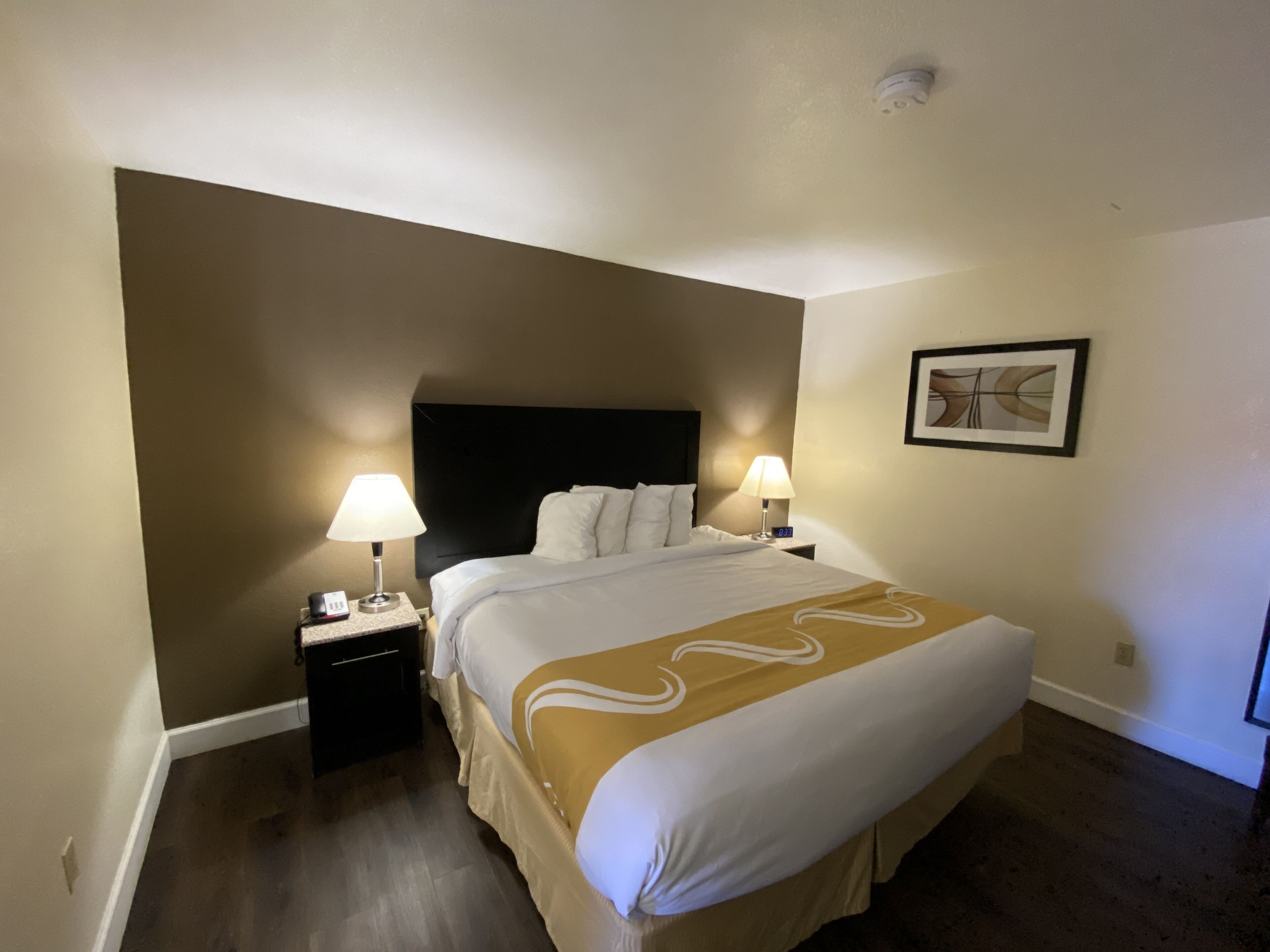 A hotel room with a bed, two bedside tables with lamps, a wall clock, and a framed picture.