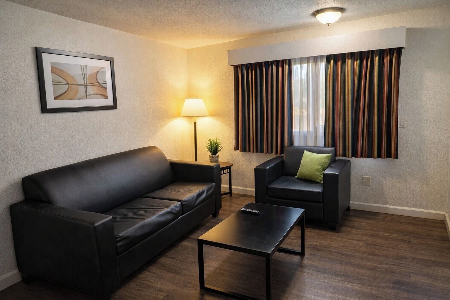 A modern living room with a black leather sofa, a matching armchair, a small table, a floor lamp, and a window with striped curtains.