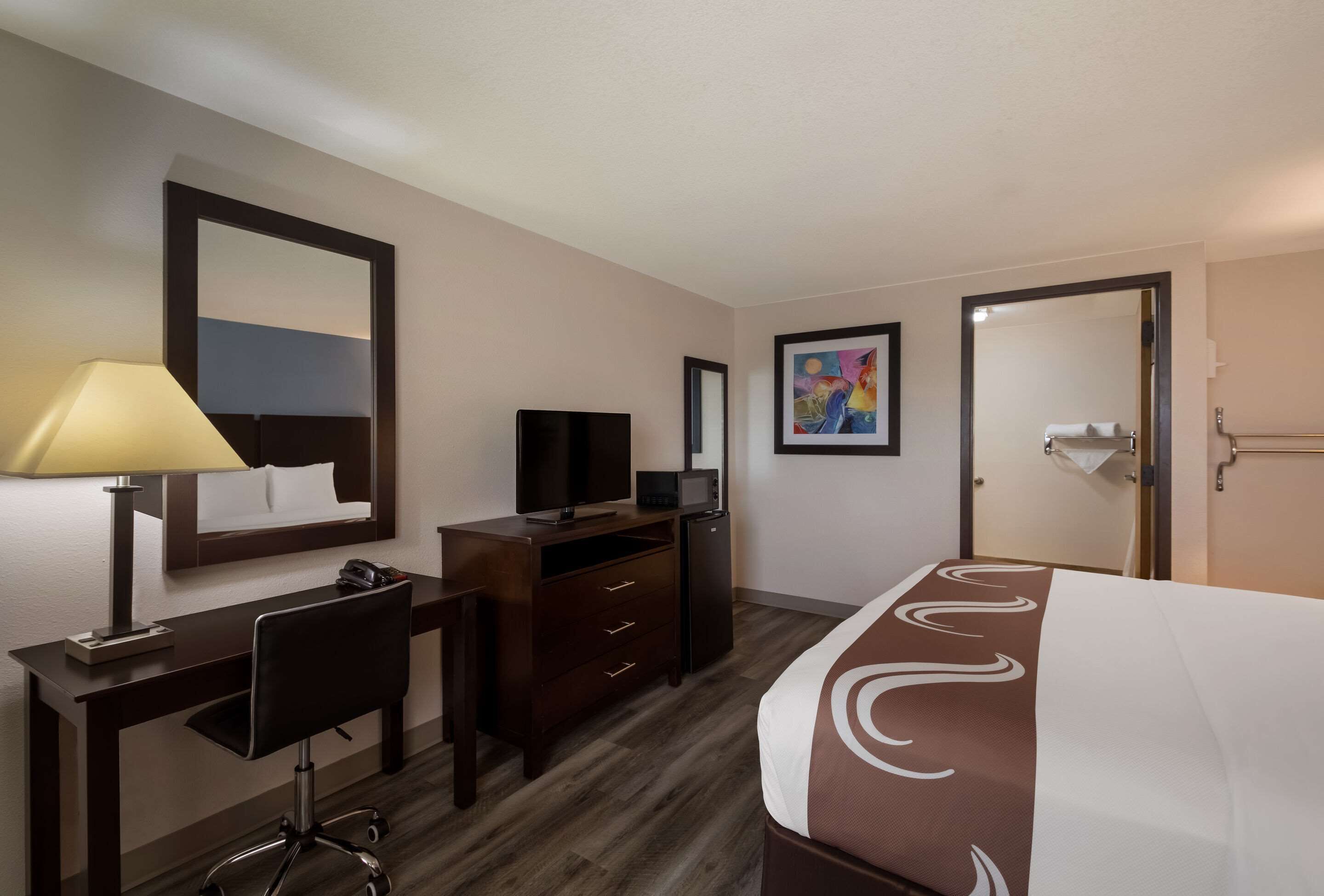 A hotel room with a bed, a desk with a lamp, a TV, a dresser, and a painting on the wall.