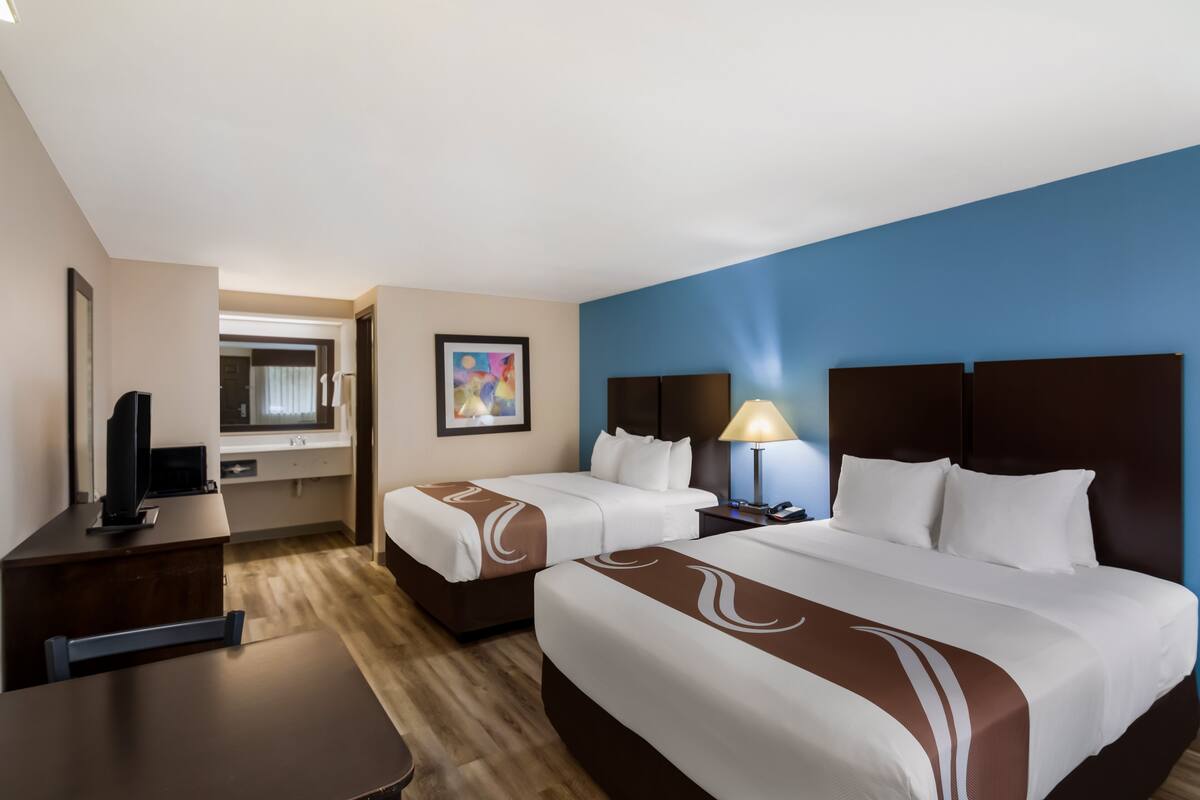 A hotel room with two beds, a desk, a TV, a lamp, and a painting on the wall.