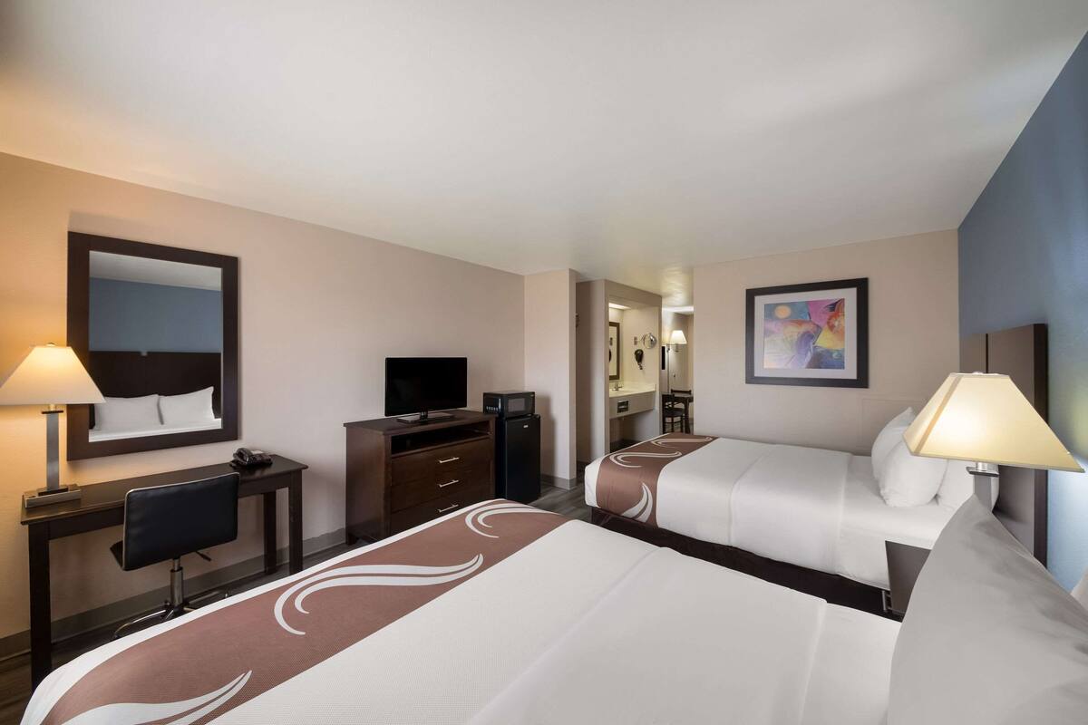 A hotel room with two beds, a desk with a chair, a television, and a mirror.