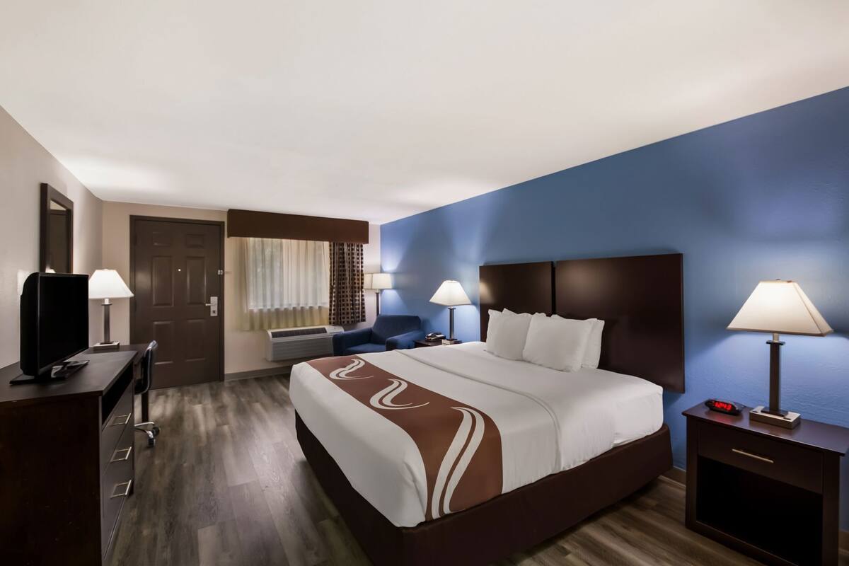 A hotel room with a bed, a desk, a chair, a TV, a lamp, and a clock.